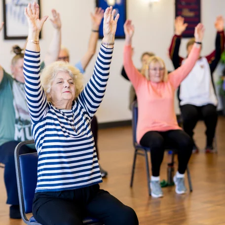 Ältere Frauen sitzen auf Stühlen und heben die Arme während einer Sportübung.