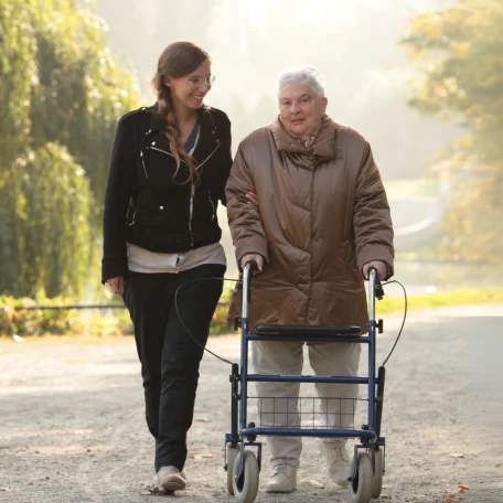 Eine Pflegerin geht mit einer älteren Dame, die einen Rollator benutzt, am Lietzensee spazieren.